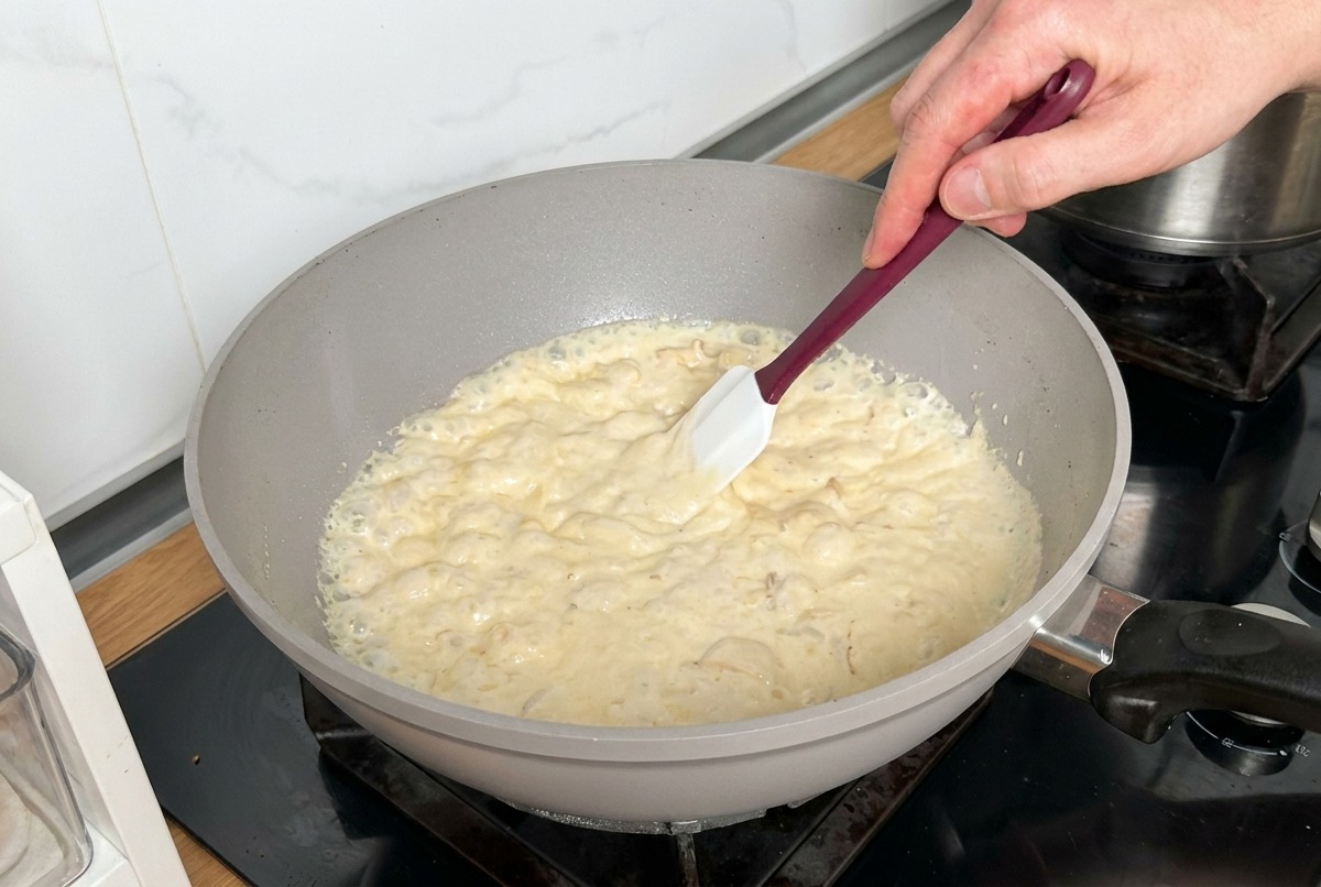 A thick, bubbling cream sauce with mushrooms and bacon being stirred in a pan with a white spatula.