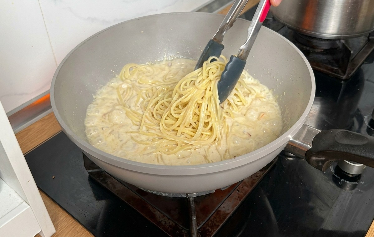 Cooked long pasta being transferred with metal tongs from a pot into a pan of creamy sauce.