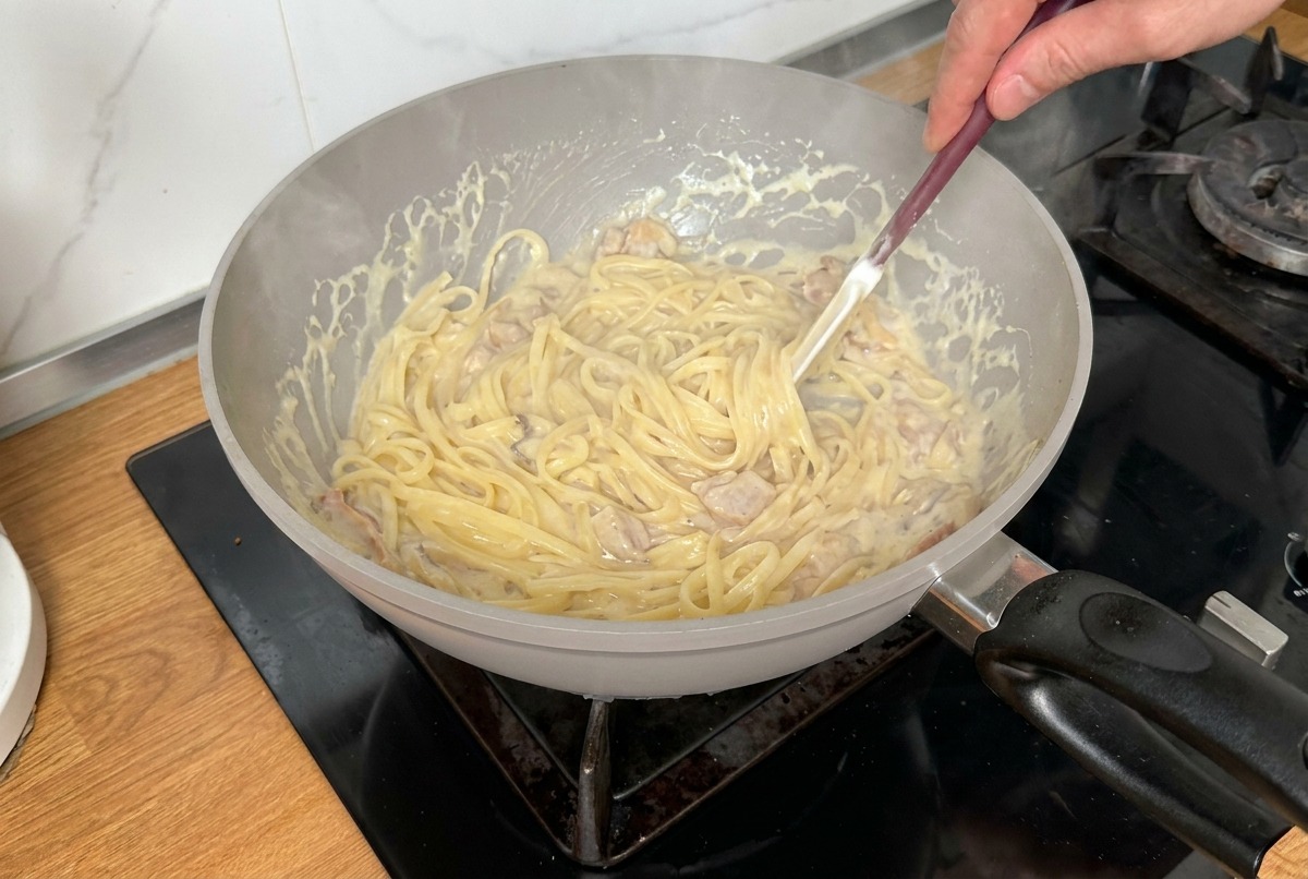 Pasta being tossed and mixed with a spatula in a creamy sauce to ensure even coating.