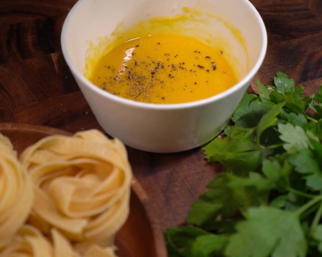 Coarse black pepper flakes sprinkled over a smooth yellow egg and cheese sauce in a white bowl.