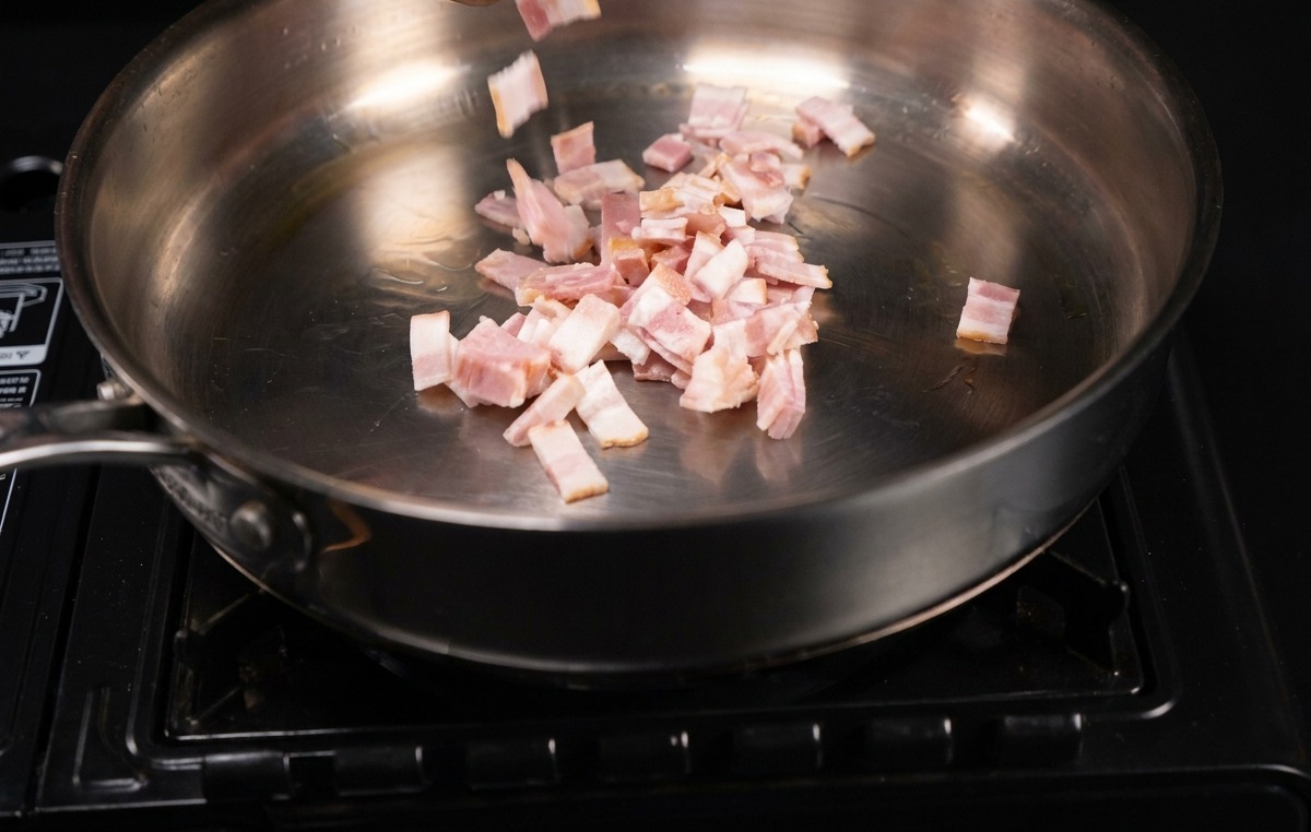 Small rectangular pieces of bacon being dropped into a shimmering metal skillet.