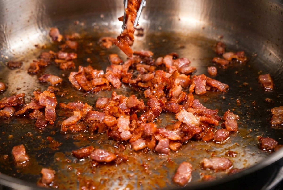 A stainless steel pan filled with small, crispy brown bacon bits being lifted out with metal tongs.