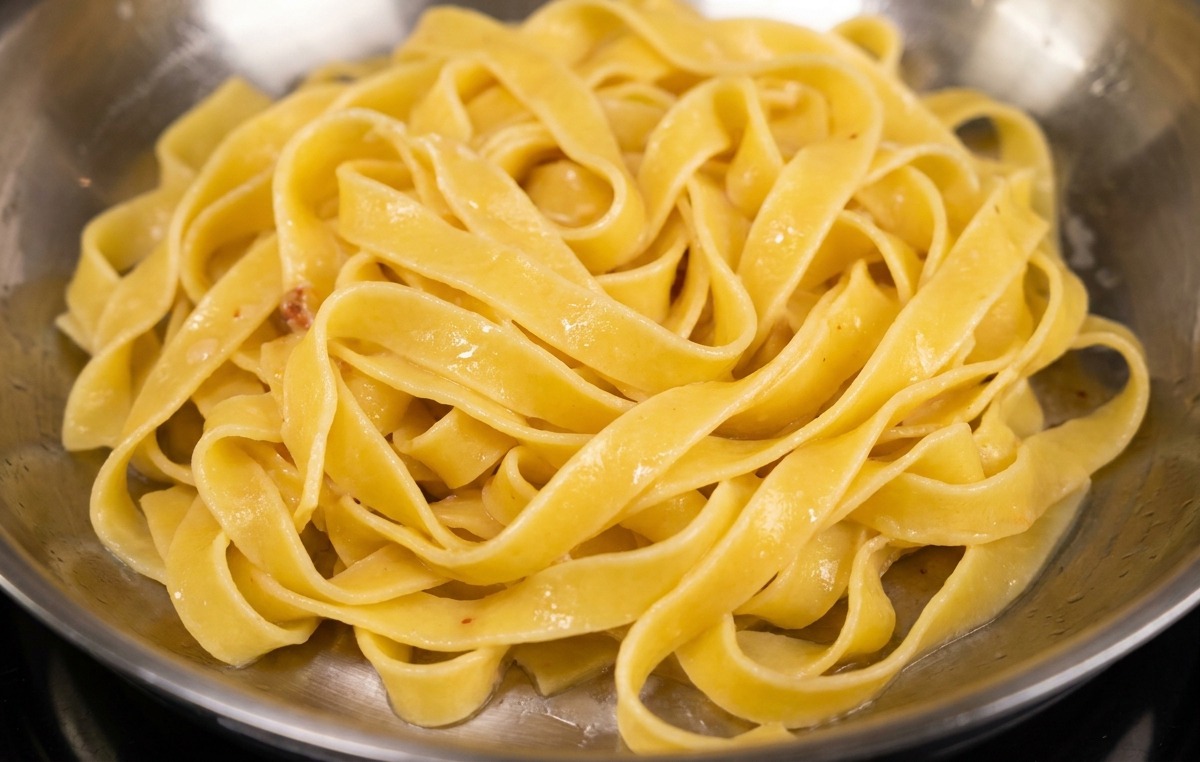 Fettuccine pasta being added to a stainless steel pan containing golden-brown fried bacon and rendered fat.