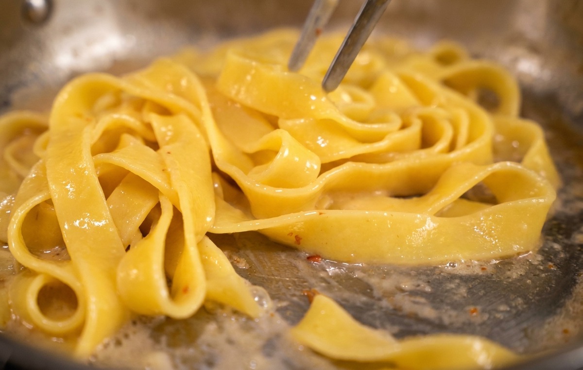 A pair of tongs tossing long pasta in a pan to emulsify a yellow egg-based sauce into a creamy coating.