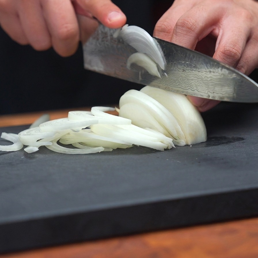 A close-up shot of a person using a sharp knife to create thin, uniform slices of white onion on a dark grey cutting board.