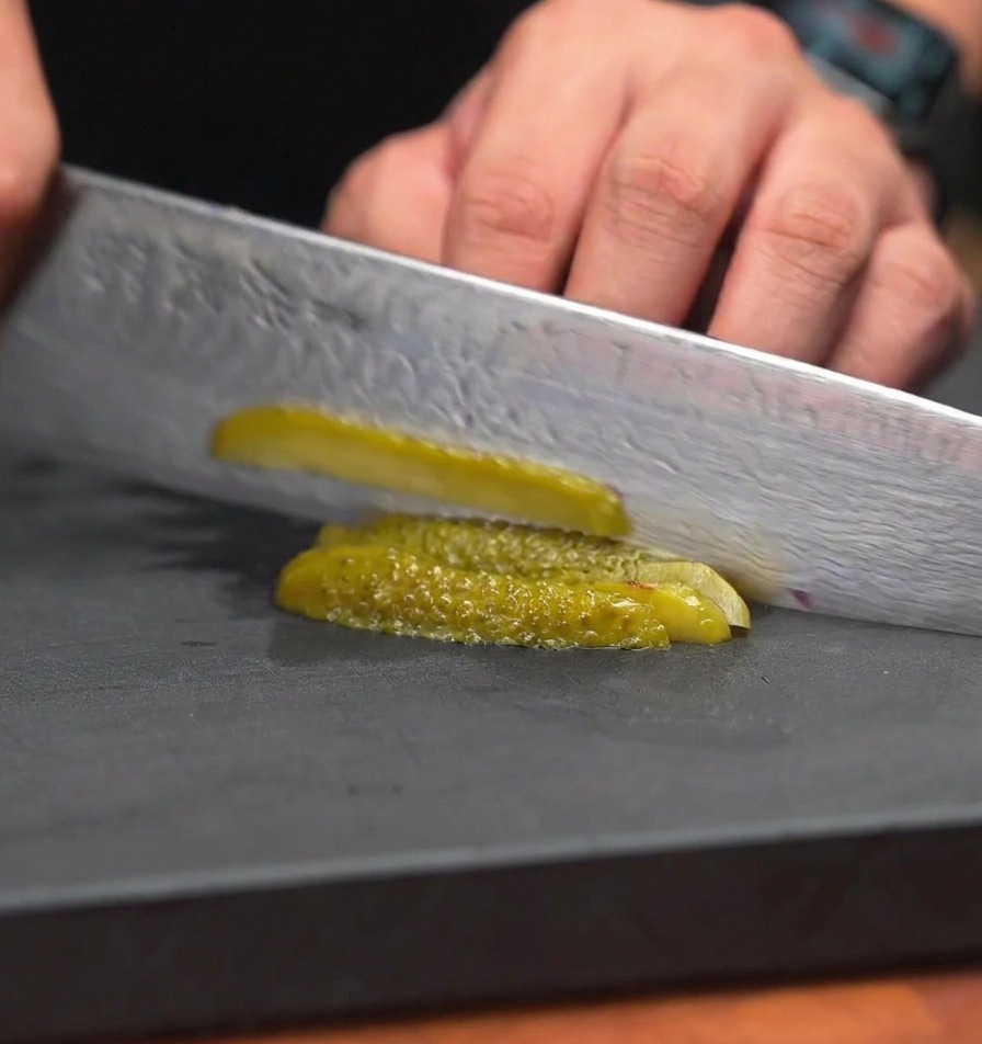 A chefs knife dicing green pickled cucumbers into very small, uniform pieces on a black work surface.