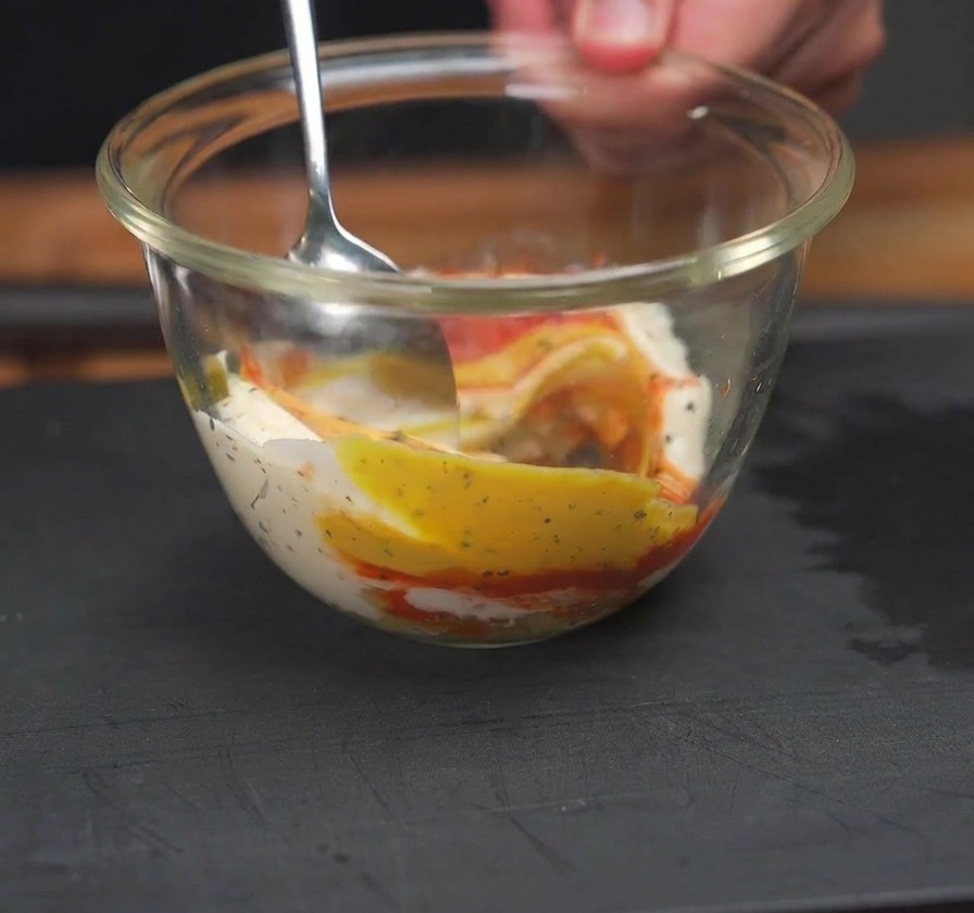 A spoon stirring together red ketchup, yellow mustard, and white mayonnaise in a clear glass bowl.
