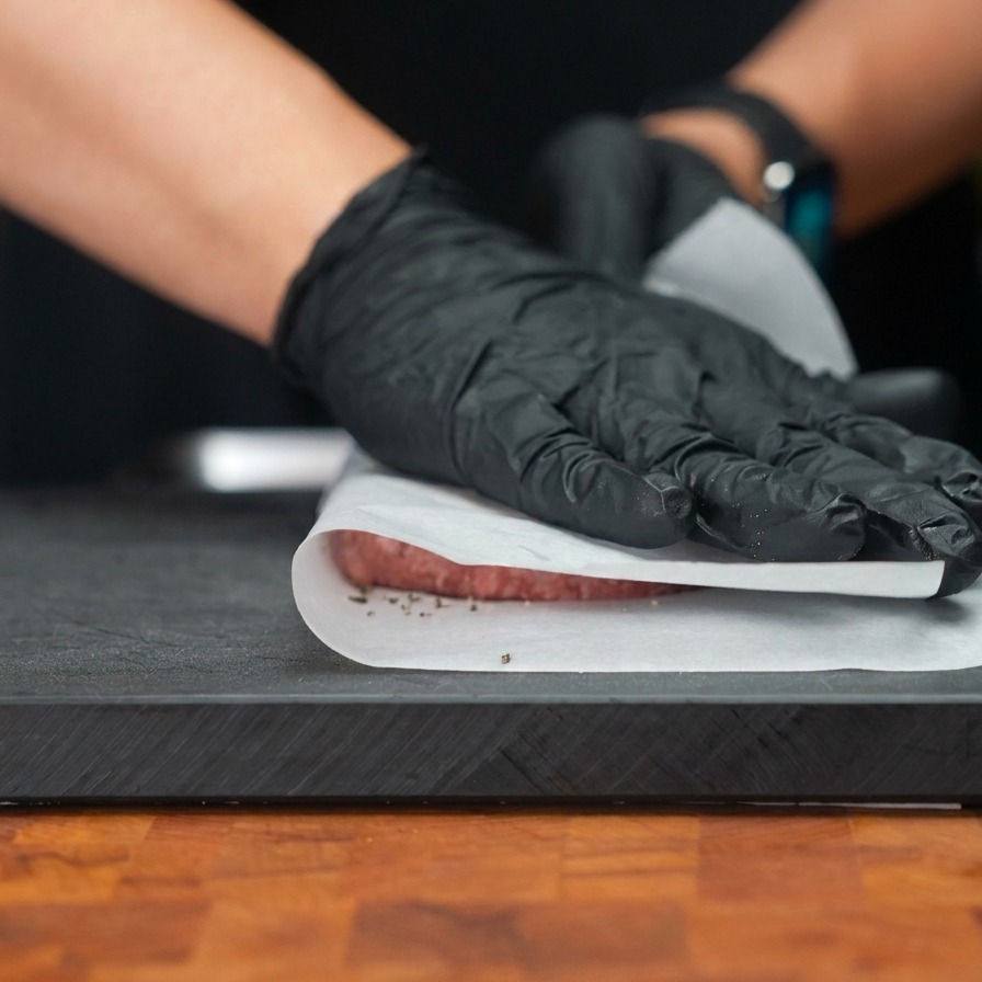 Gloved hands pressing down on a beef patty through a layer of parchment paper to flatten it into a wide, thin disk.
