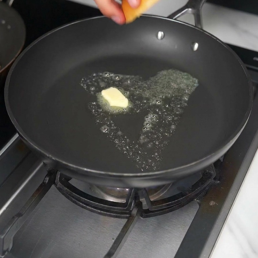 A hand placing a brioche burger bun into a frying pan with a melting cube of yellow butter.
