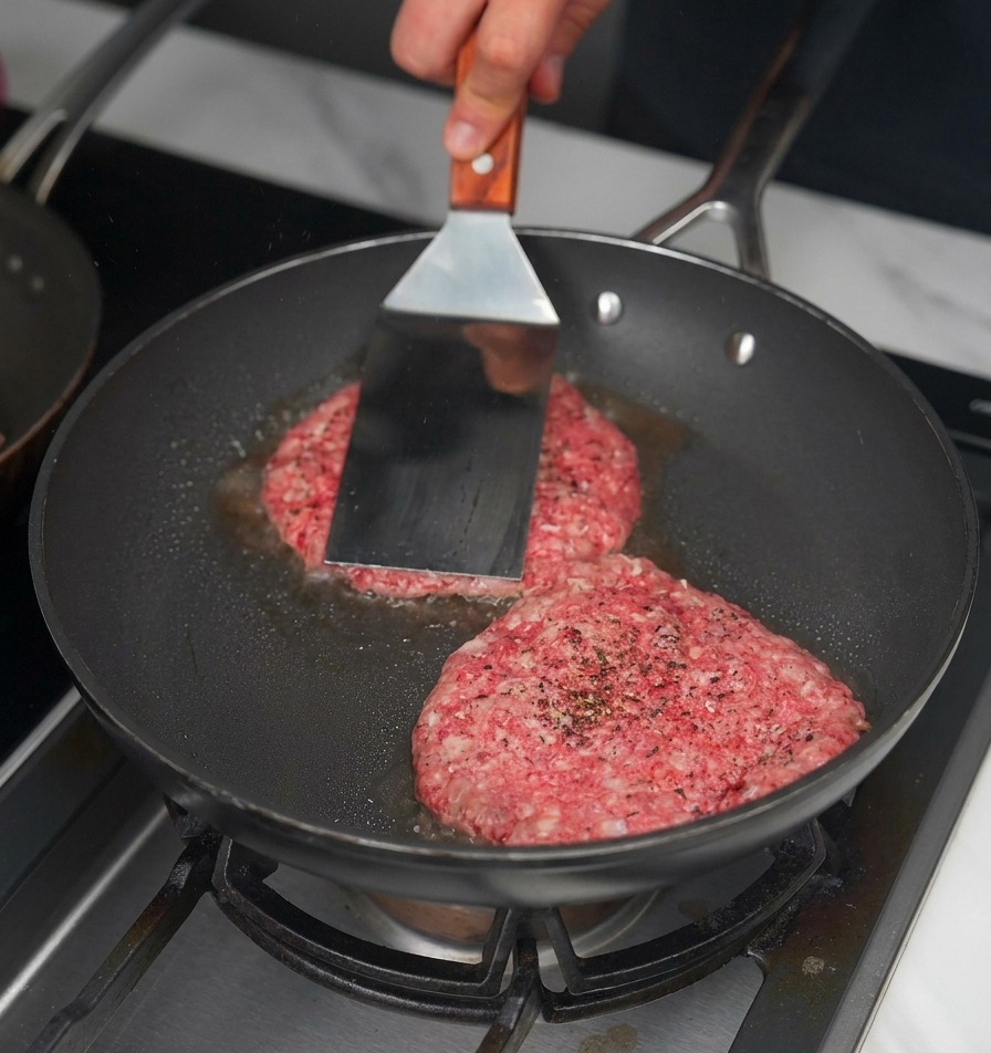 A metal spatula pressing down on two raw beef patties in a black frying pan to create a crust.