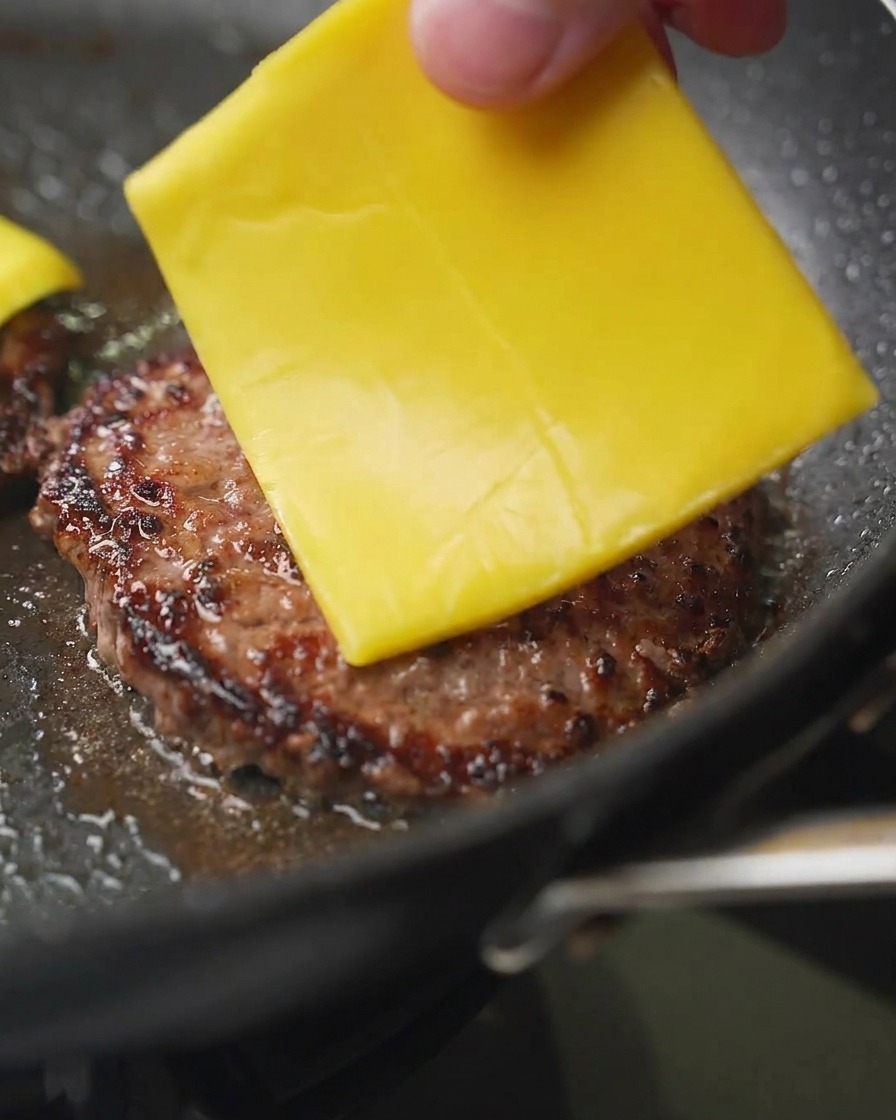 A close-up shot of a hand placing a square slice of yellow cheddar cheese onto a browned beef patty in a pan.