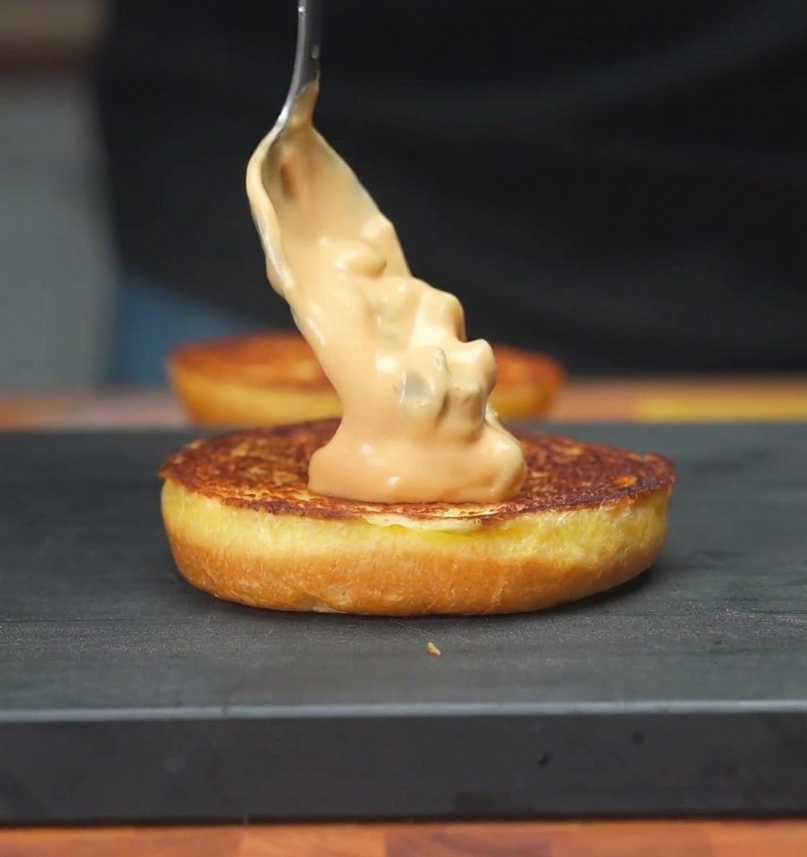 A spoon spreading a creamy orange-colored pickle sauce onto the toasted surface of a burger bun.