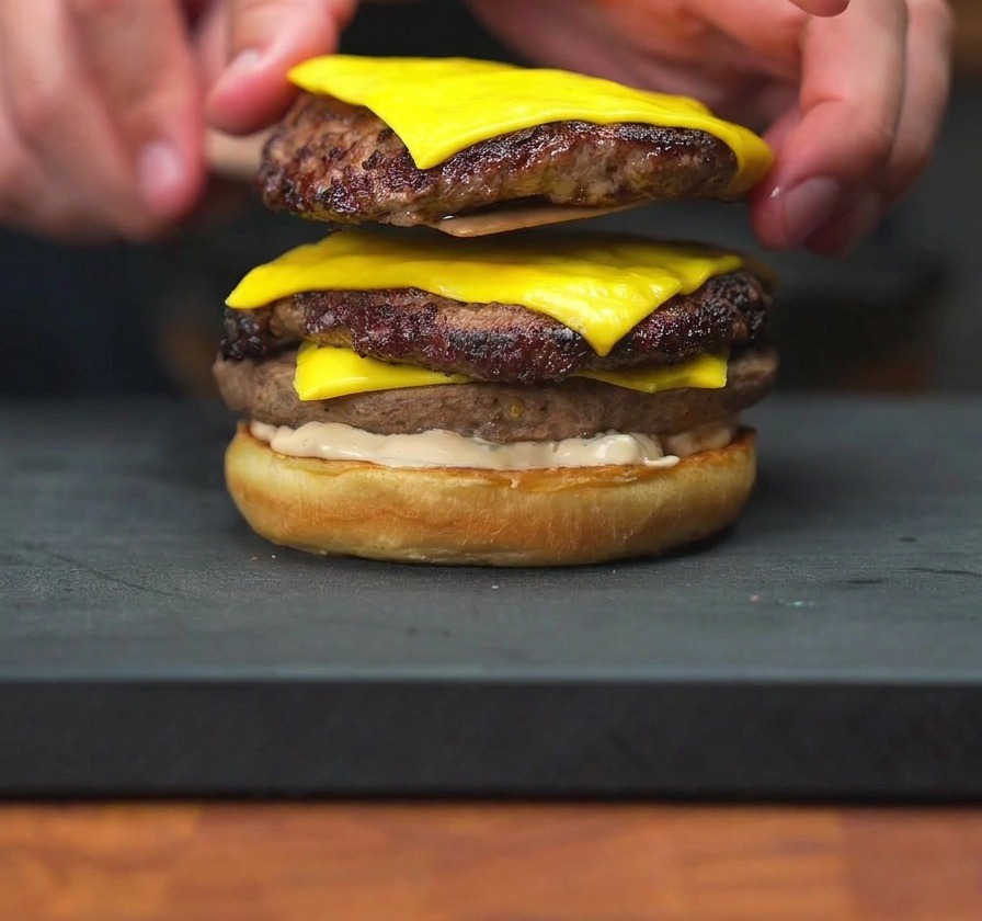 A hand lowering a stack of two cheeseburger patties with melted cheese onto a sauced burger bun.