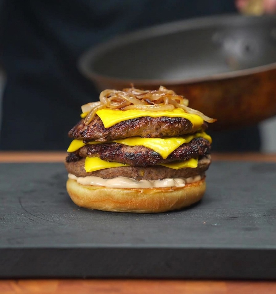 Caramelized onions being placed onto a stack of cheeseburger patties on a bun using a pair of chopsticks.
