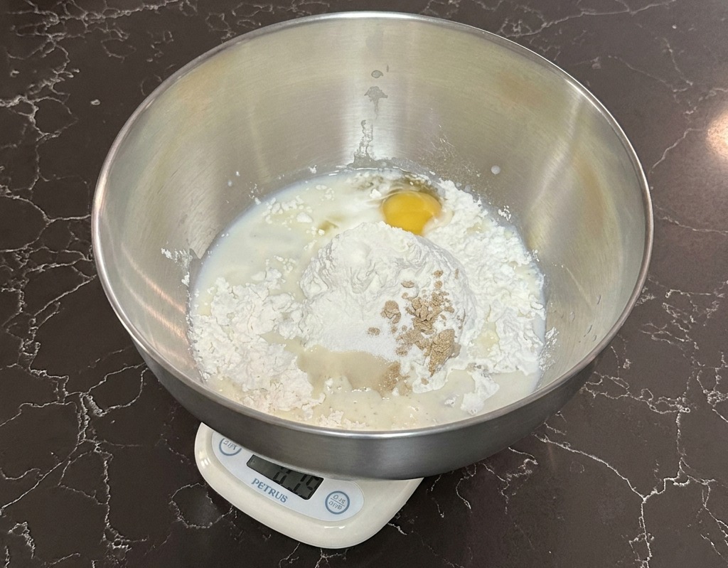 A stainless steel mixing bowl sitting on a digital scale containing flour, milk, an egg, and yeast.