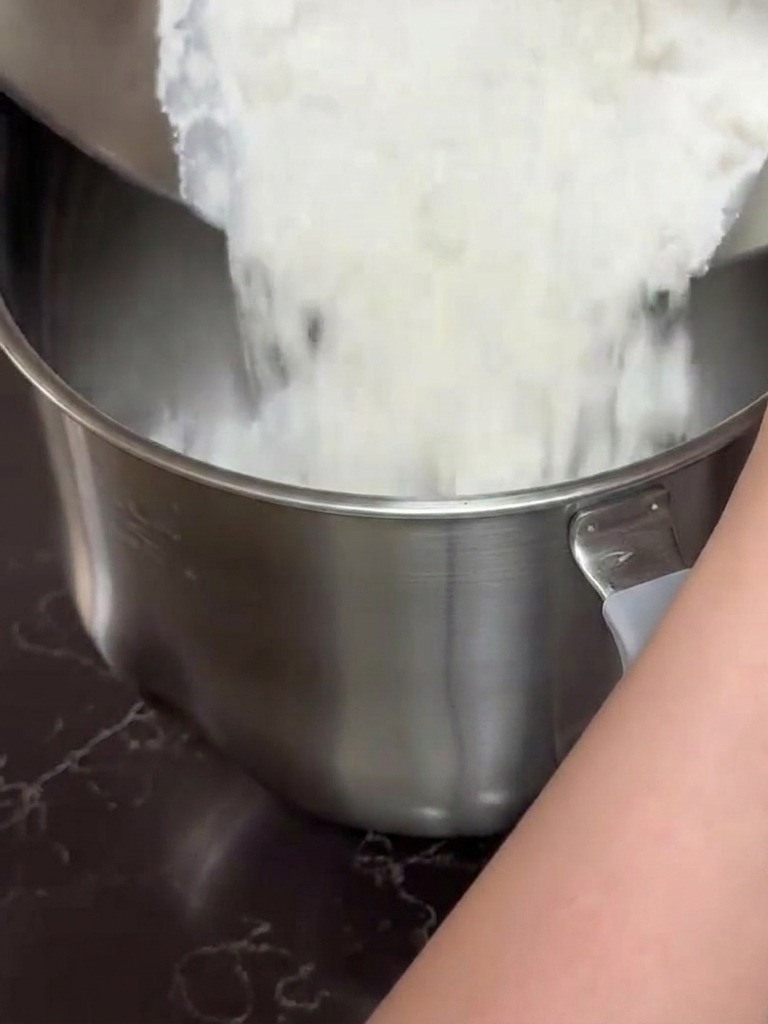 Hands pouring a white flour and milk mixture from a stainless steel bowl into a large stand mixer bowl.