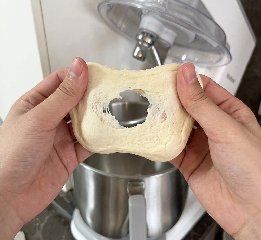A person stretching a piece of pale dough between their hands to check for the formation of a gluten membrane.
