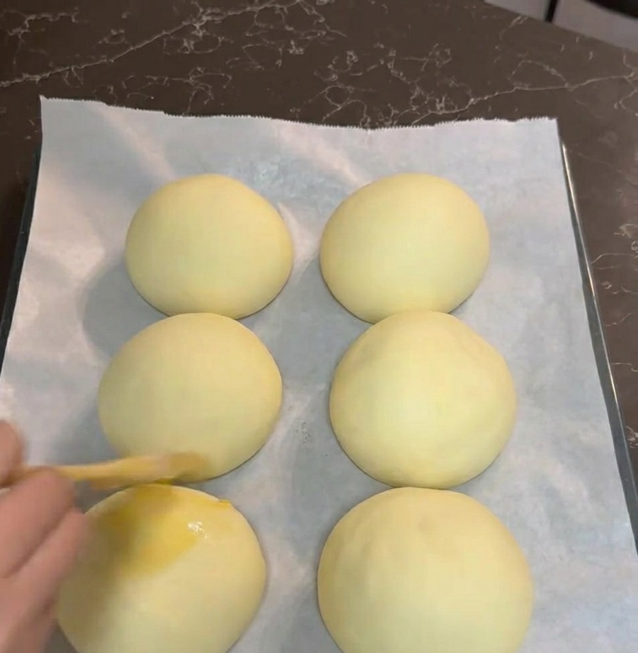 A hand using a pastry brush to coat six smooth, proofed dough balls with a yellow egg wash on a parchment-lined tray.