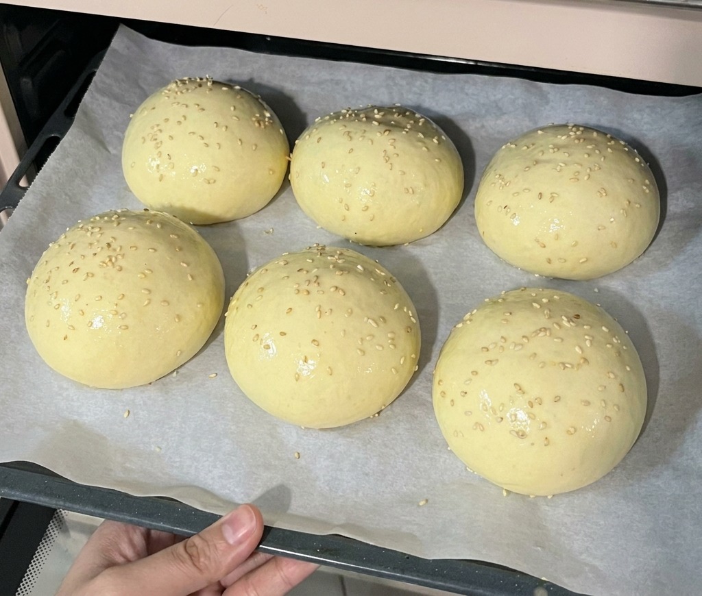 A baking tray holding six proofed burger buns topped with sesame seeds being placed into a preheated oven.