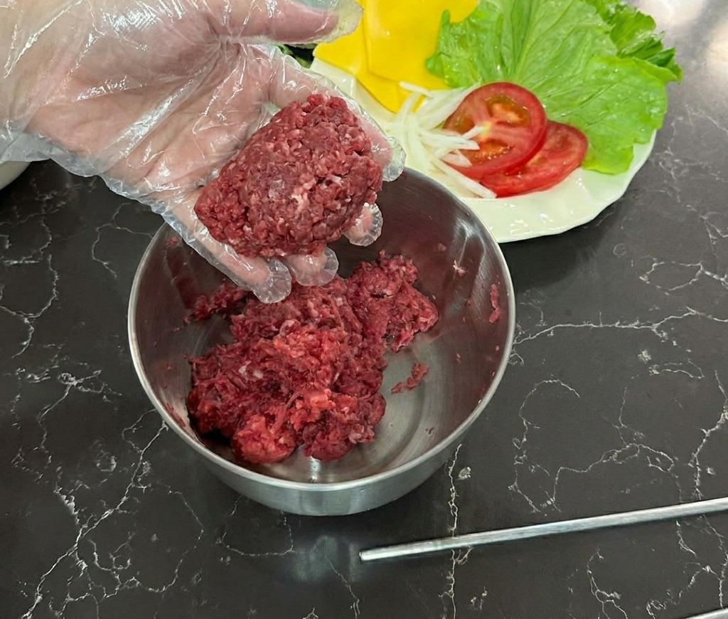 A hand in a clear plastic glove holding a rounded ball of raw ground beef over a stainless steel mixing bowl.