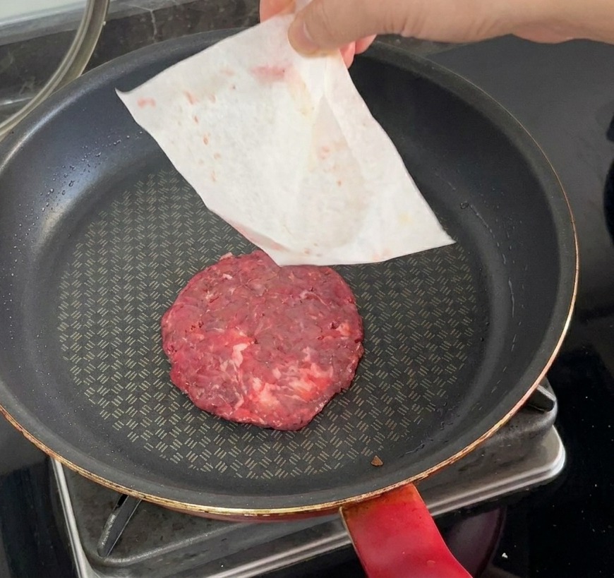 A raw beef patty being placed into a hot skillet with the parchment paper being peeled away by hand.