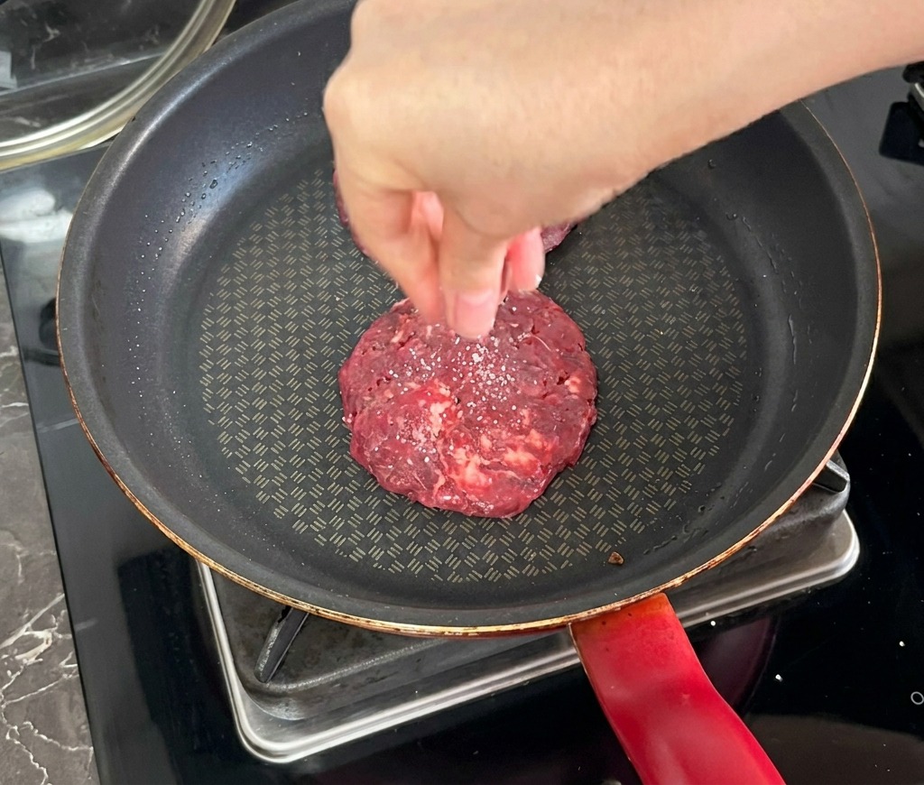 A hand sprinkling salt over a beef patty that is being seared in a dark non-stick frying pan.