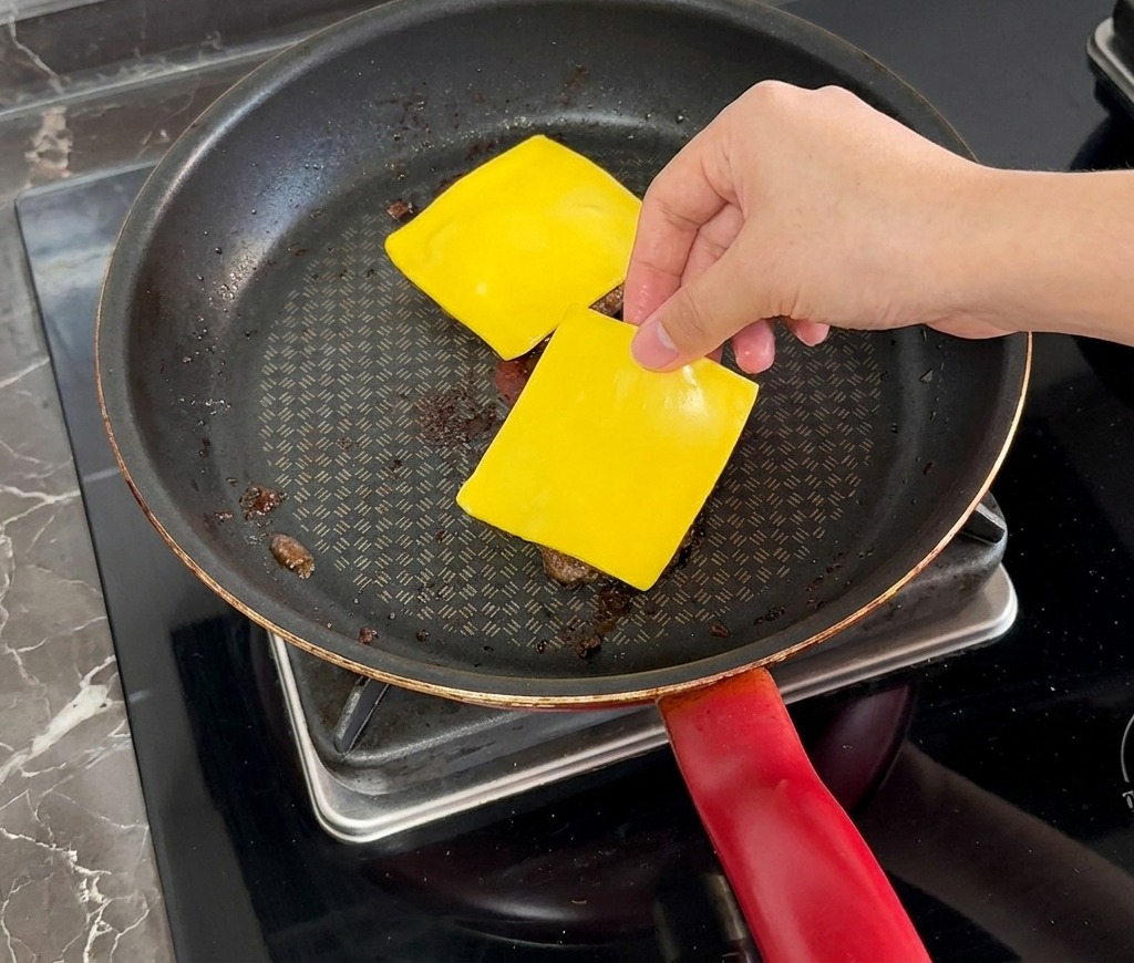 A hand placing a square slice of yellow cheese onto a cooked beef patty in a pan.