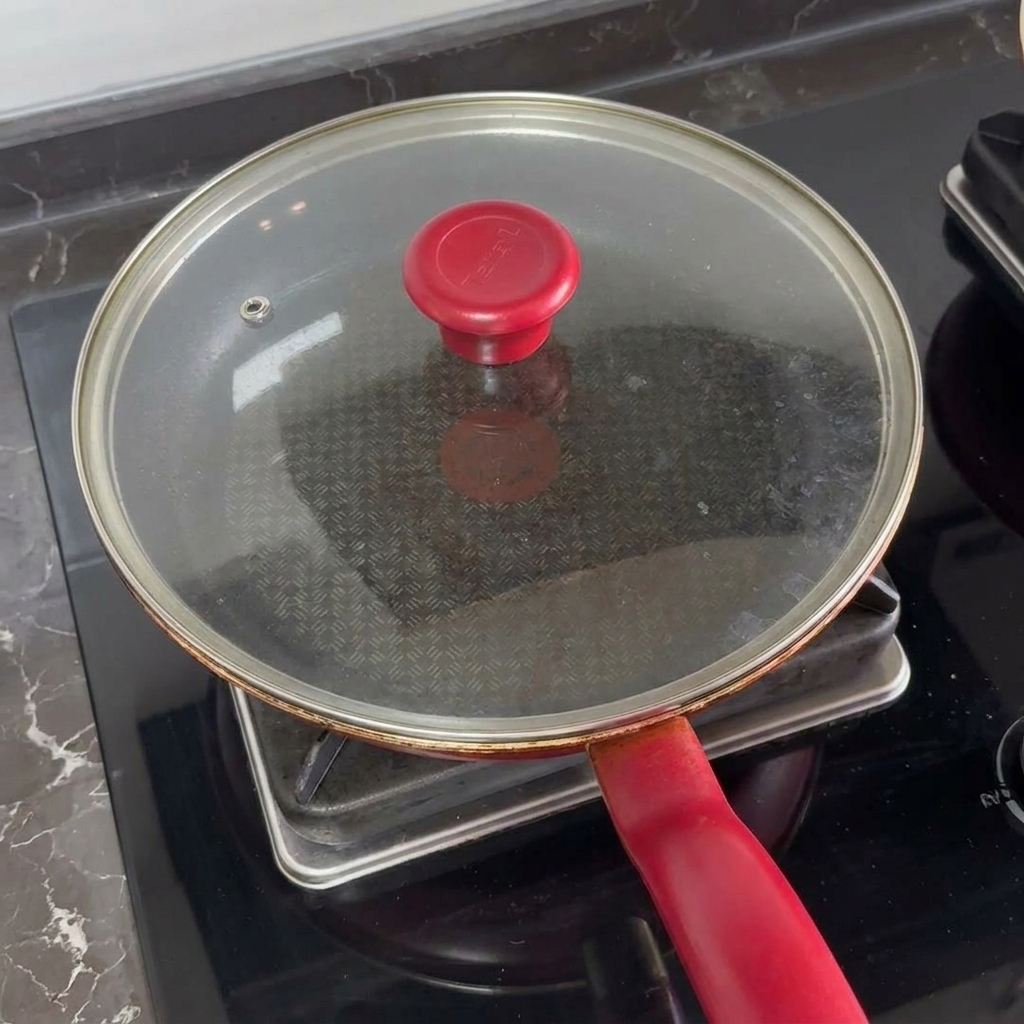 A glass lid with a red handle covering a frying pan on a stovetop to melt cheese over burger patties.