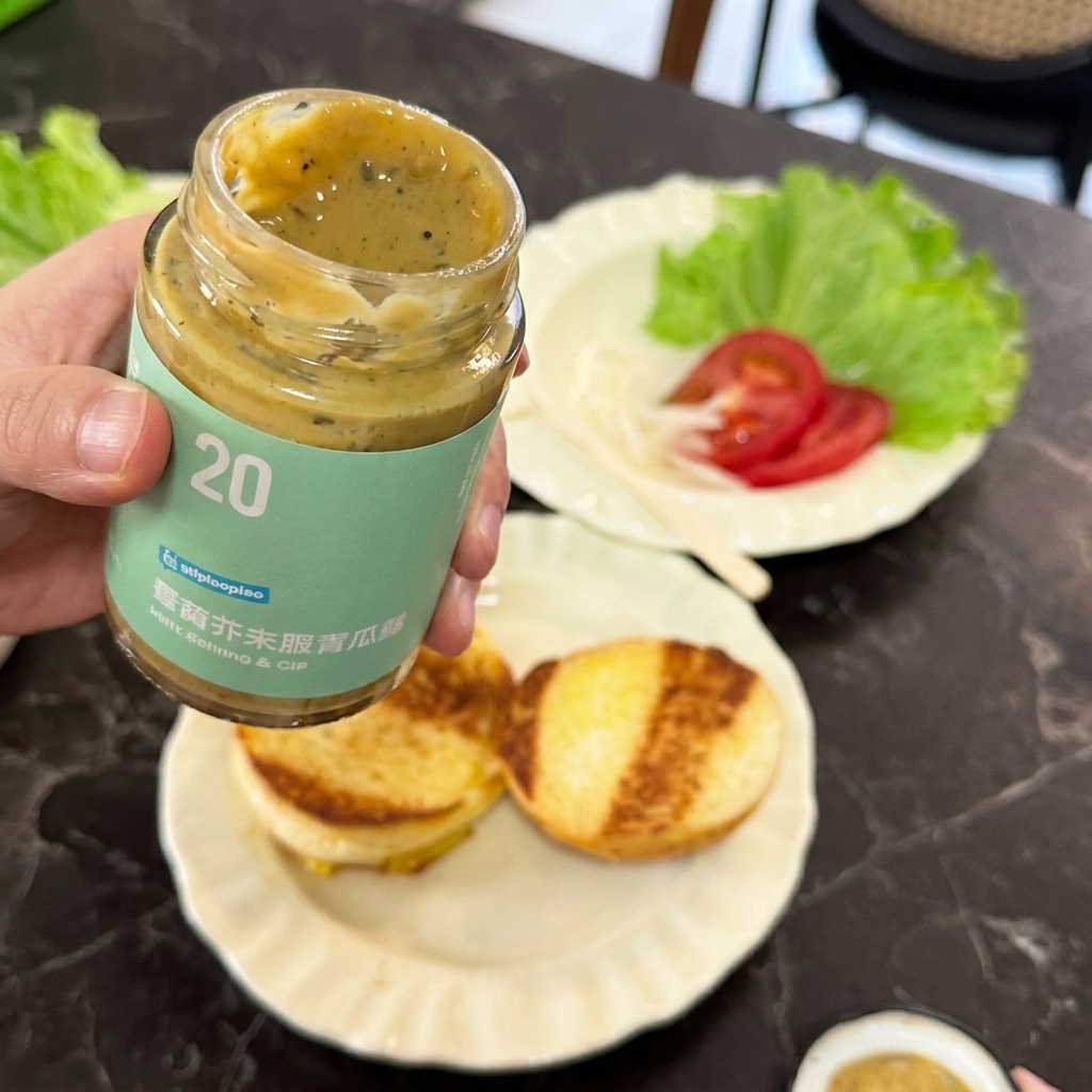 A hand holding an open jar of mustard pickle sauce over a plate of golden toasted burger buns.