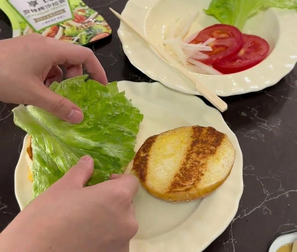 Hands placing a fresh green lettuce leaf onto a toasted burger bun on a white scalloped plate.