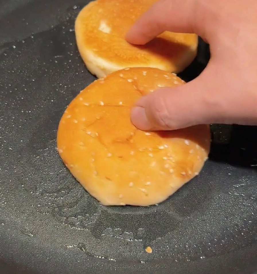 Halved burger buns being toasted cut-side down in a dry frying pan until the surfaces are golden and toasted.
