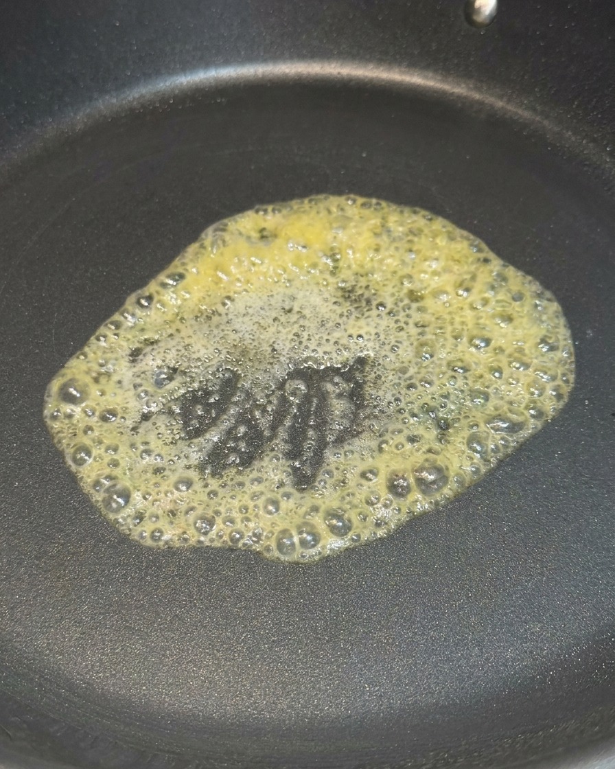 A knob of yellow butter melting and foaming in the center of a black non-stick skillet over heat.
