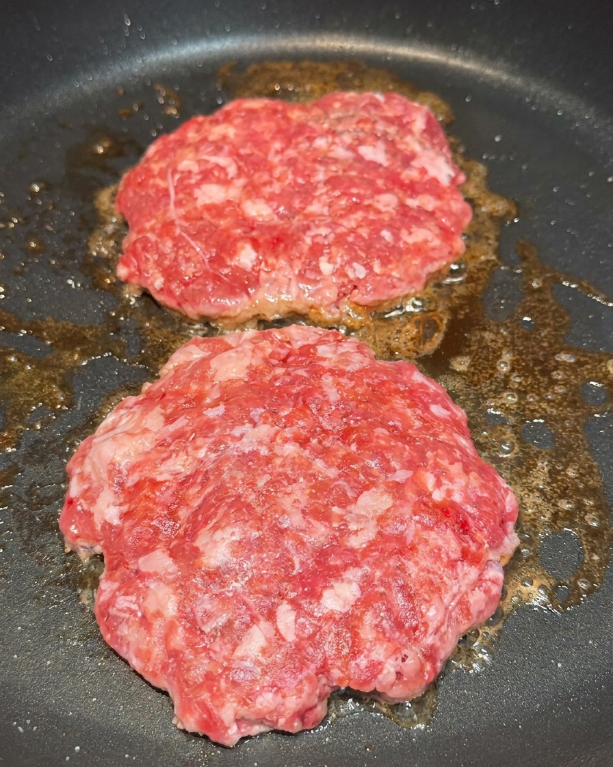 Two raw beef patties in a frying pan being seasoned with visible grains of salt and black pepper.