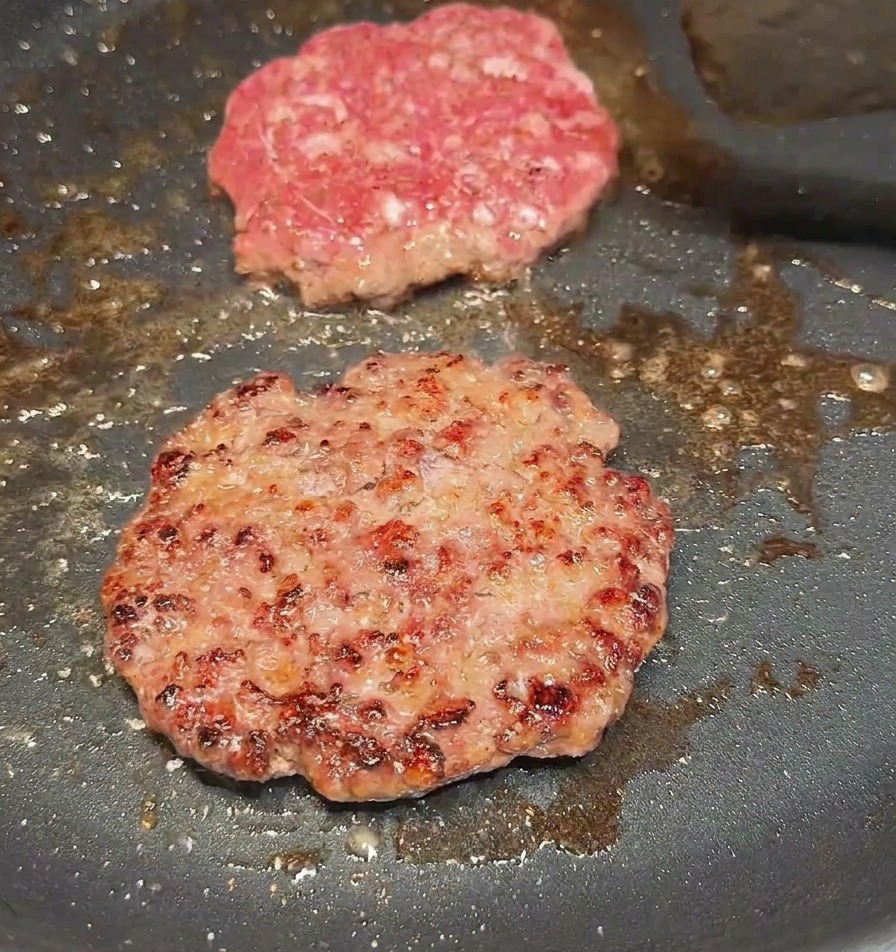 A spatula flipping a beef patty in a pan to reveal a well-browned, seared crust on the cooked side.