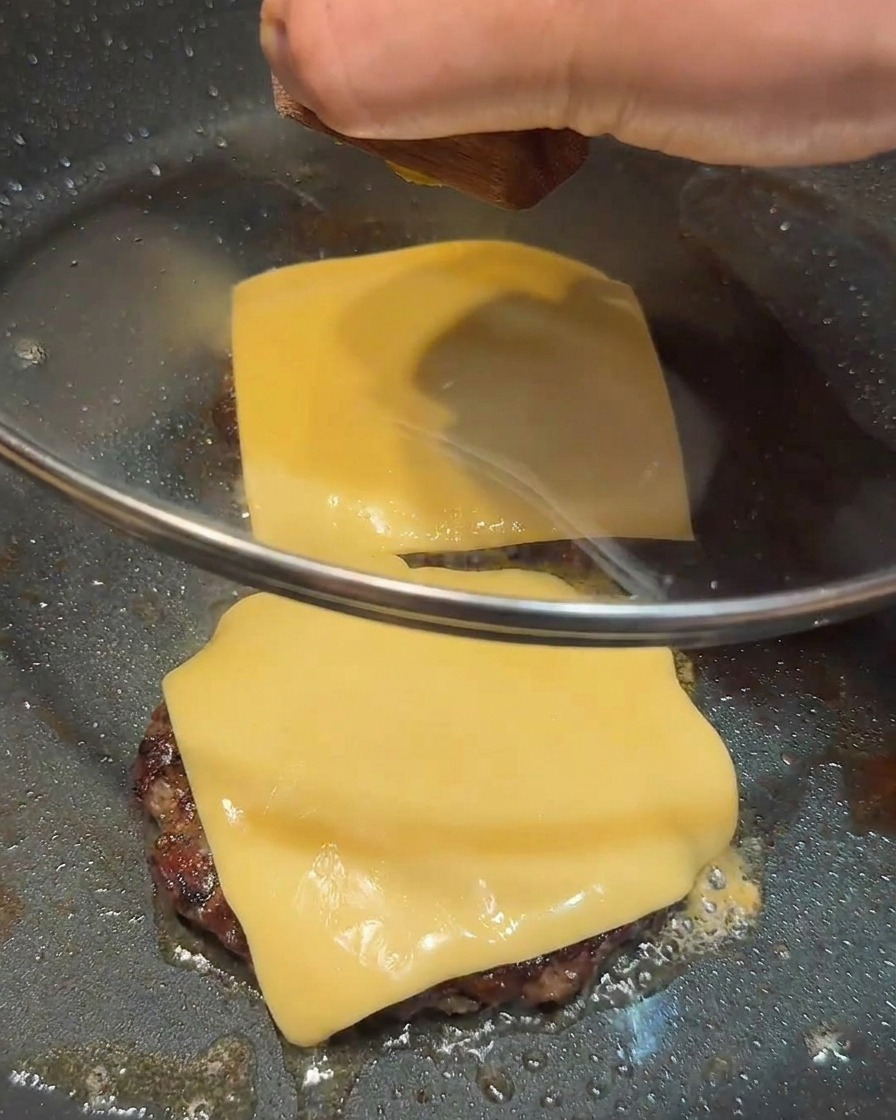 A glass lid covering a pan containing two beef patties topped with square slices of yellow cheese.