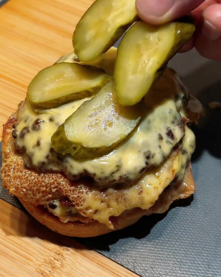 Fingers arranging slices of green pickles on top of a double beef patty stacked with melted cheese.