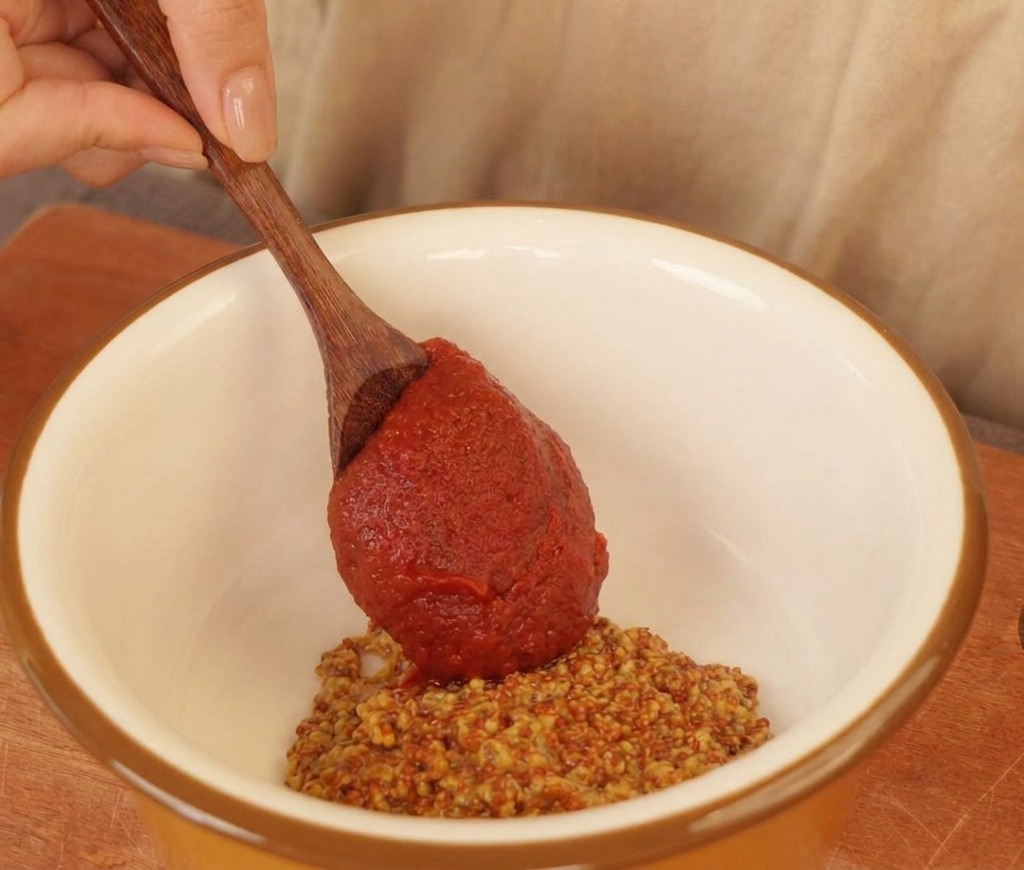 A wooden spoon adding thick red chili sauce to a white bowl containing whole-grain mustard and mayonnaise.