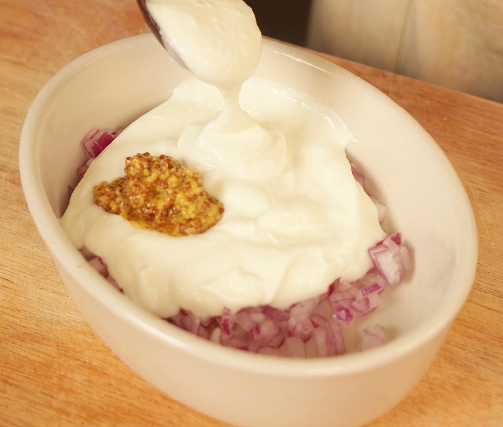 A spoonful of creamy white yogurt being added to a small oval dish containing chopped red onions and grainy mustard.