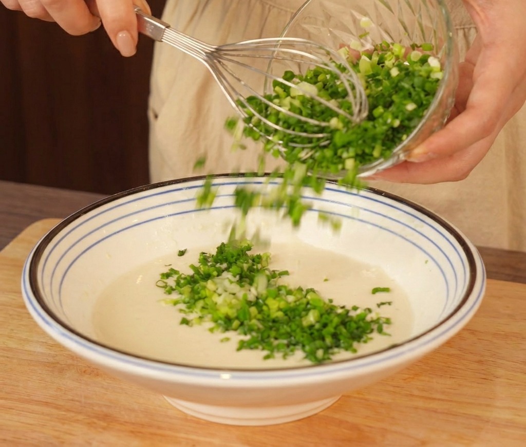 A large bowl of pale batter being mixed with a whisk as a generous pile of chopped green scallions is added.