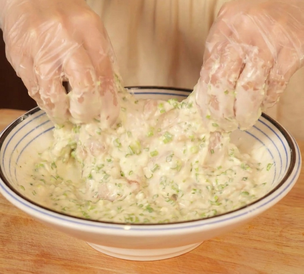 Gloved hands mixing chicken thighs in a bowl of thick creamy white batter filled with chopped green scallions.