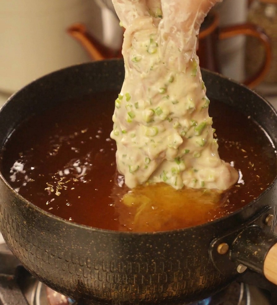 A hand in a clear plastic glove lowering a piece of chicken heavily coated in scallion batter into a deep pot of hot frying oil.