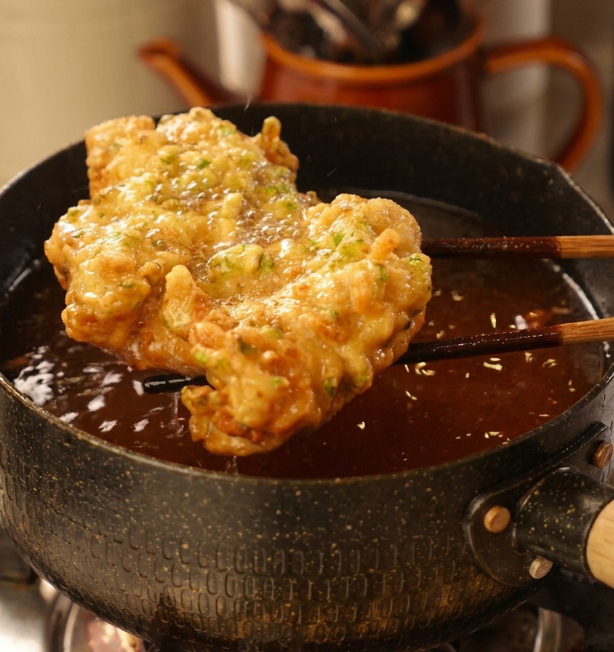 Large, golden-brown crispy fried chicken being lifted out of a pot of oil with dark wooden chopsticks.