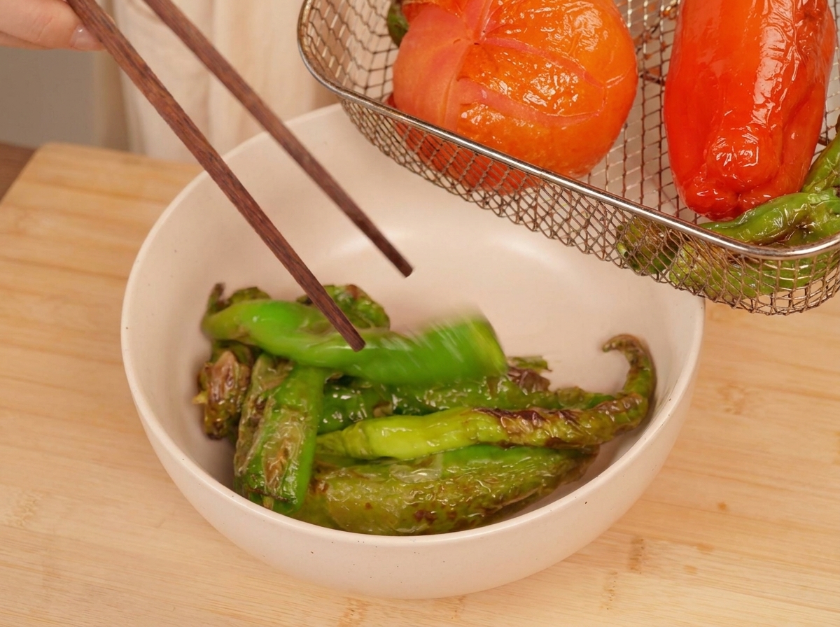 Wooden chopsticks picking up a roasted green pepper to place it in a bowl, with the charred tomato visible in the air fryer basket above.