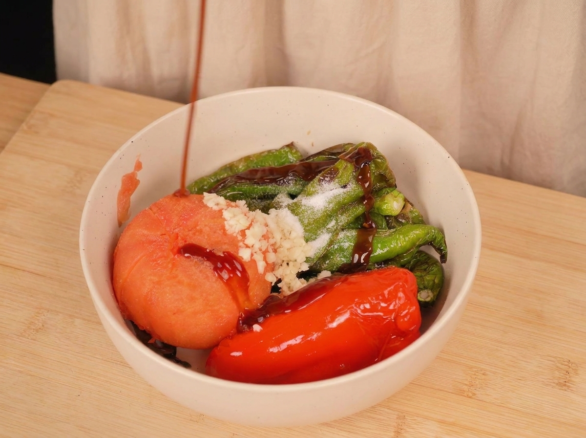 Dark oyster sauce being poured over roasted peppers, a tomato, minced garlic, and salt in a white bowl.