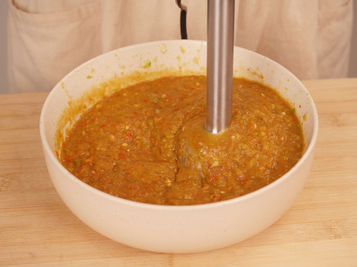An immersion blender being used inside a white bowl to blend roasted vegetables into a thick, textured orange-green sauce.