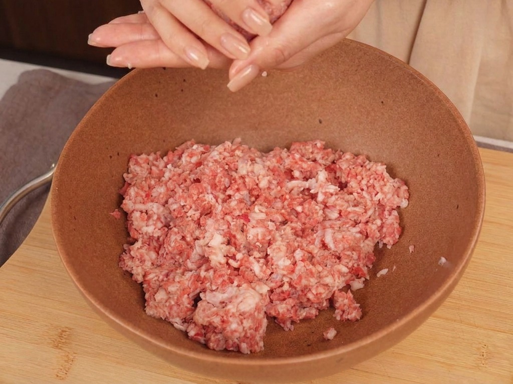 A persons hands shaping a round ball of raw ground beef above a ceramic mixing bowl.