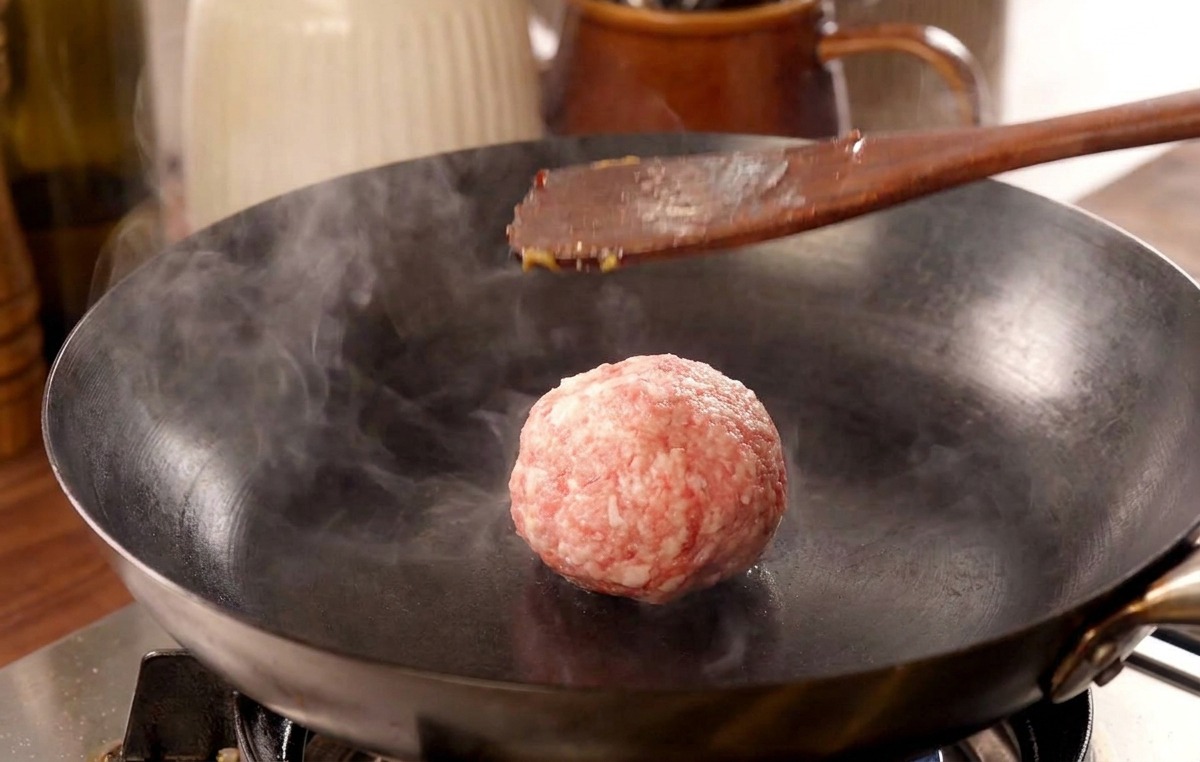 A wooden spatula pressing down on a beef meatball in a hot pan, releasing a cloud of steam.