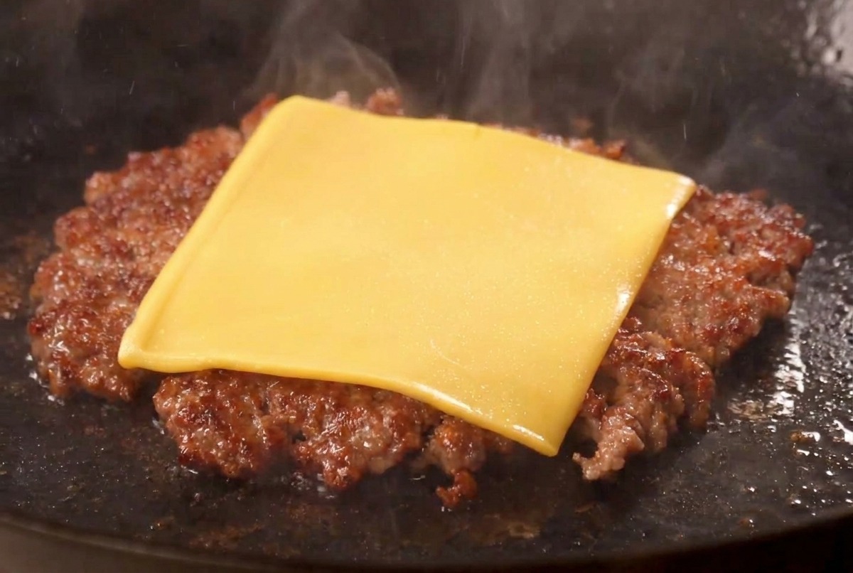 A slice of yellow American cheese melting over the top of a seared, crusted beef patty in a pan.