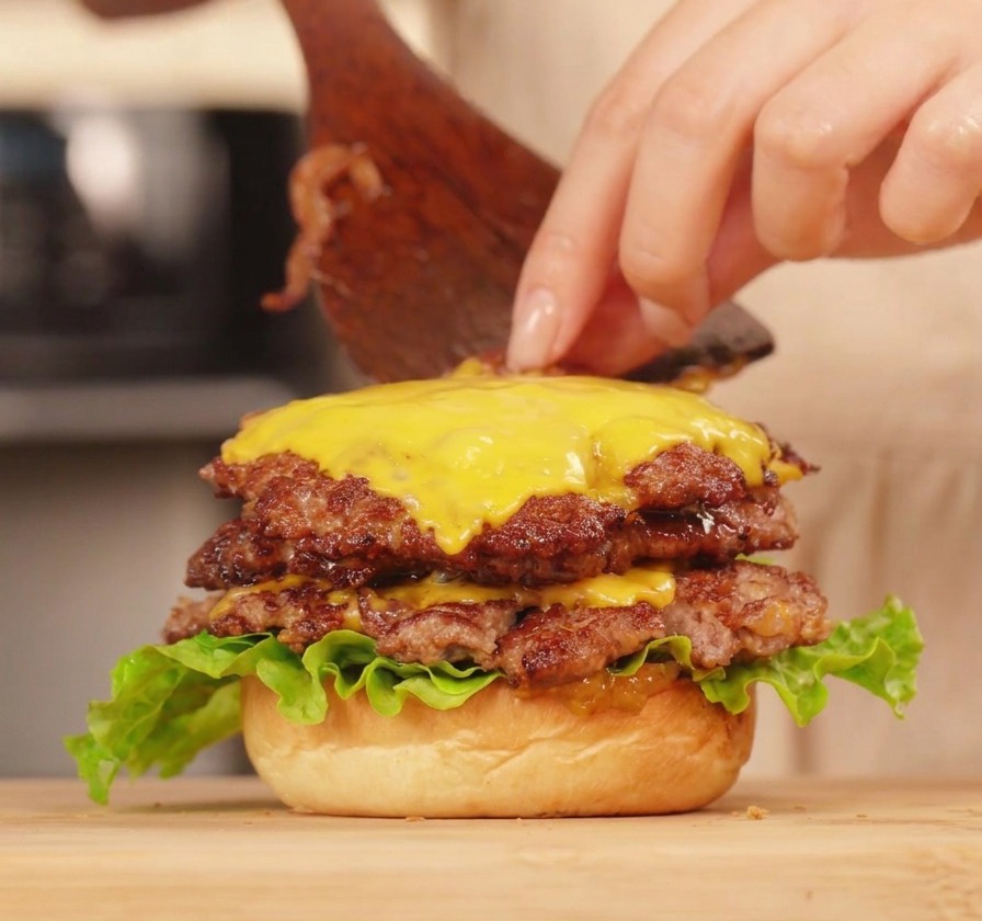 A person using a wooden spatula to assemble a double cheeseburger, applying a layer of textured green pepper sauce over melted cheese.