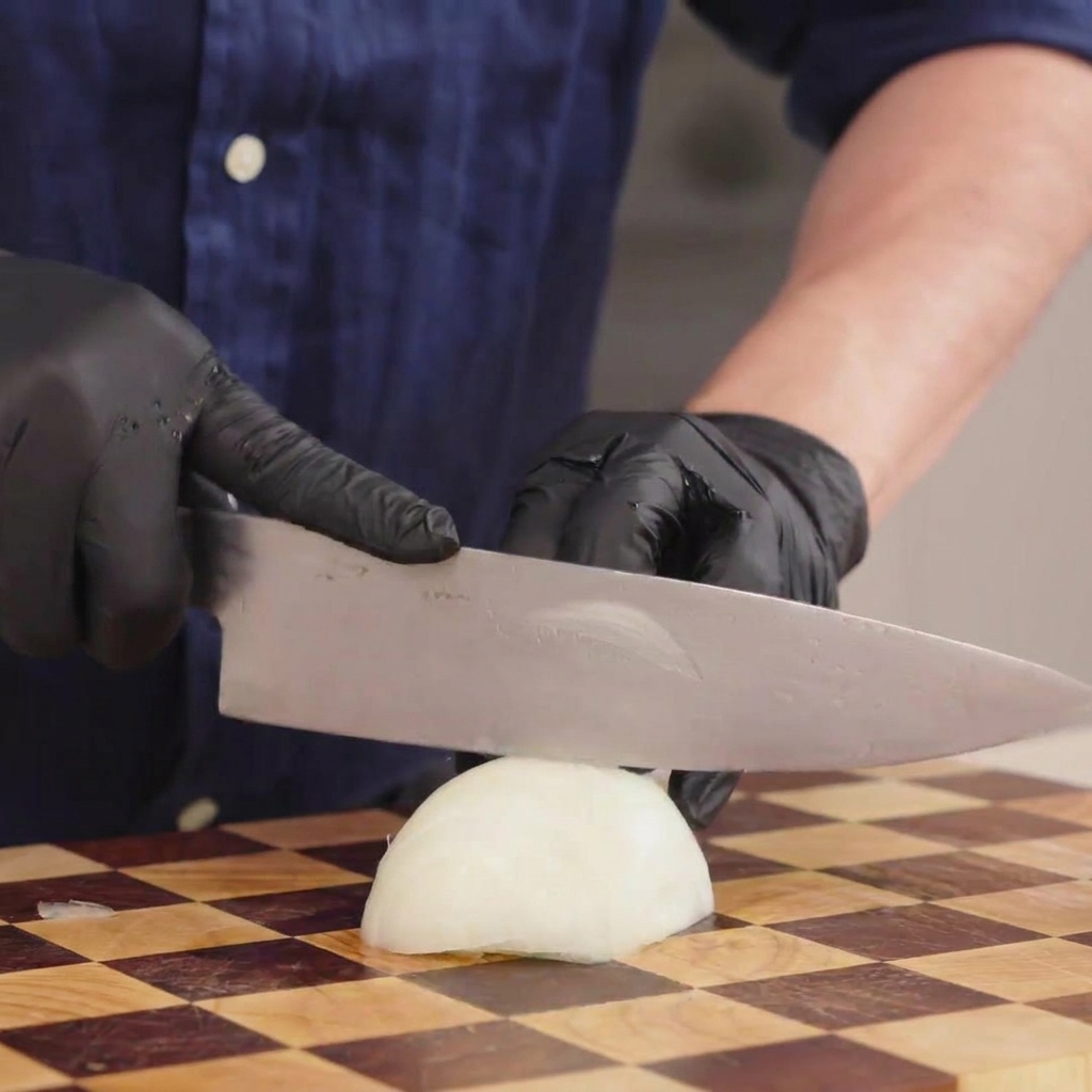 A close-up shot of a person in black gloves using a chefs knife to thinly slice a white onion on a checkered wooden board.
